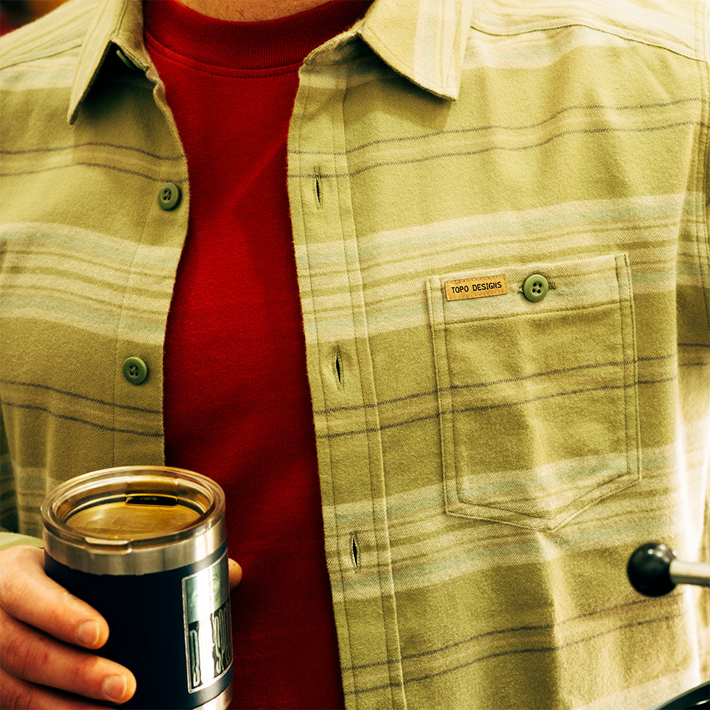 General lifestyle image of person wearing a "Oil Green Horizontal Stripe" Mountain Shirt Lightweight with a red sweater underneath, holding a black insulated mug.